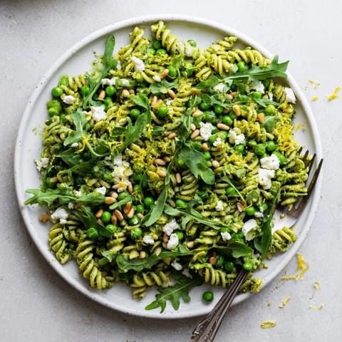 A vibrant bowl of Spring Green Pesto Pasta Salad with peas, arugula, and toasted pine nuts, served on a picnic blanket.  